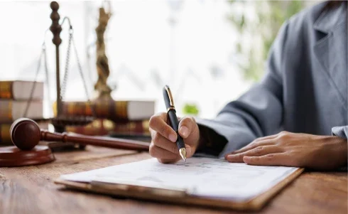 Person in a suit writing on a document at a desk with a gavel and scales of justice in the background.