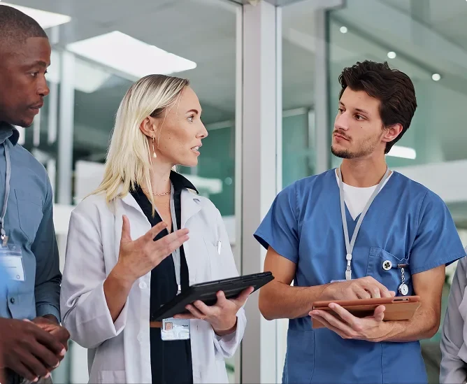A group of diverse medical professionals, including a woman in a white lab coat and a man in blue scrubs, holding tablets and engaged in a serious discussion in a modern hospital hallway