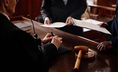 Judge holding a pen, two lawyers exchanging documents in a courtroom with a gavel on the desk.